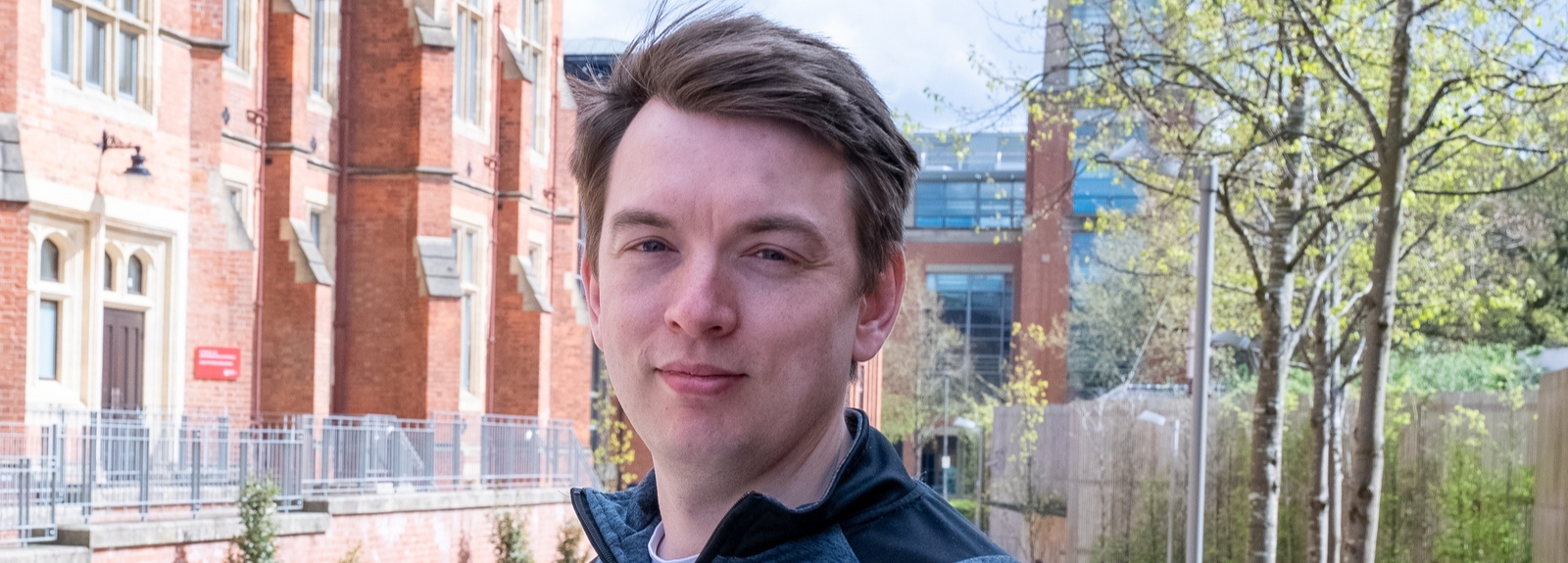 A person stands outdoors on a university campus, with red-brick buildings and trees in the background.